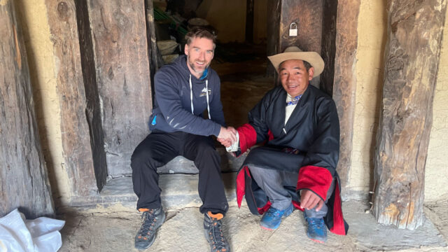 A Tetra Tech employee shaking hands with a Nepal resident - A man in casual clothing is shaking hands with a man in traditional attire, both sitting at the entrance of a rustic building.