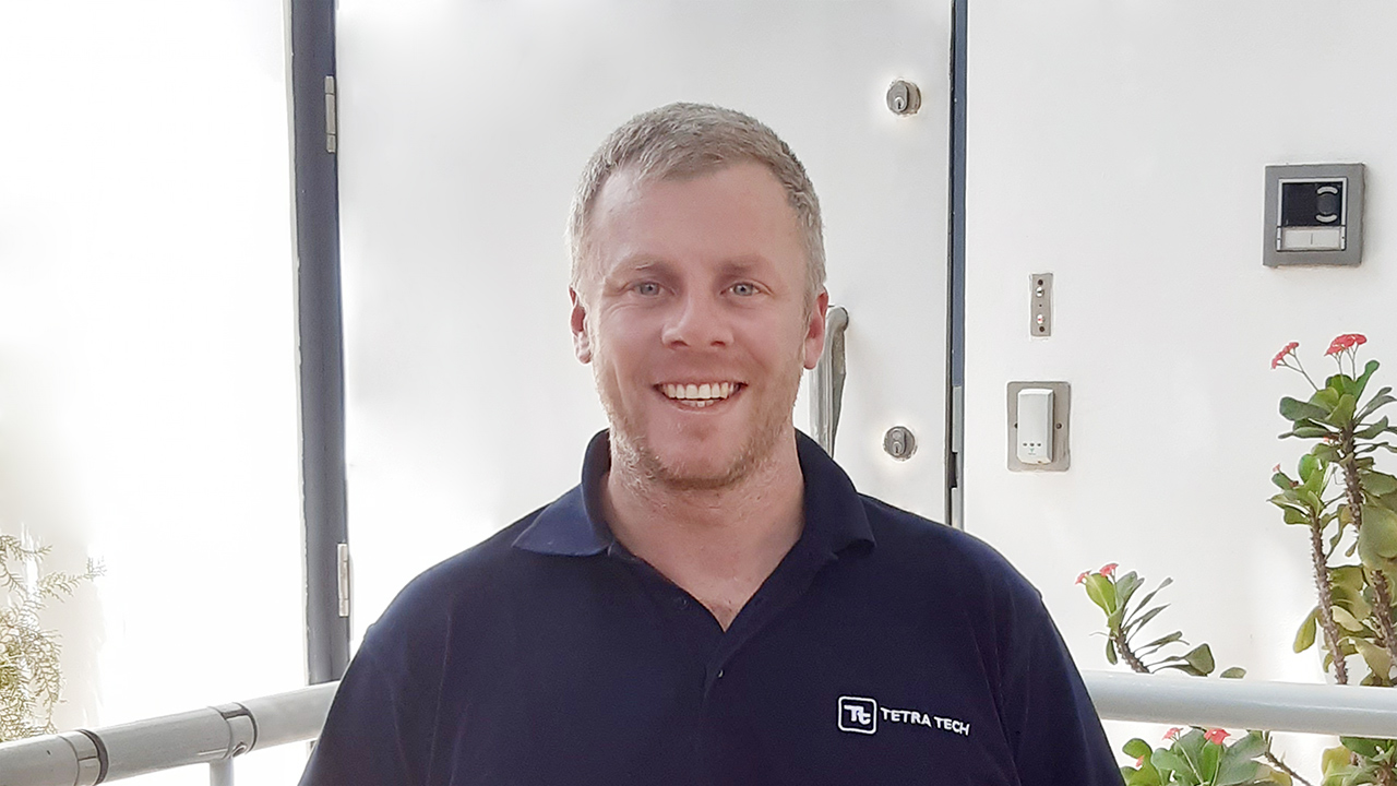 Headshot of Chris Mackowski - A smiling man wearing a navy blue T-shirt with the Tetra Tech logo stands in front of a doorway. He has short hair and a light beard, with plants visible in the background.