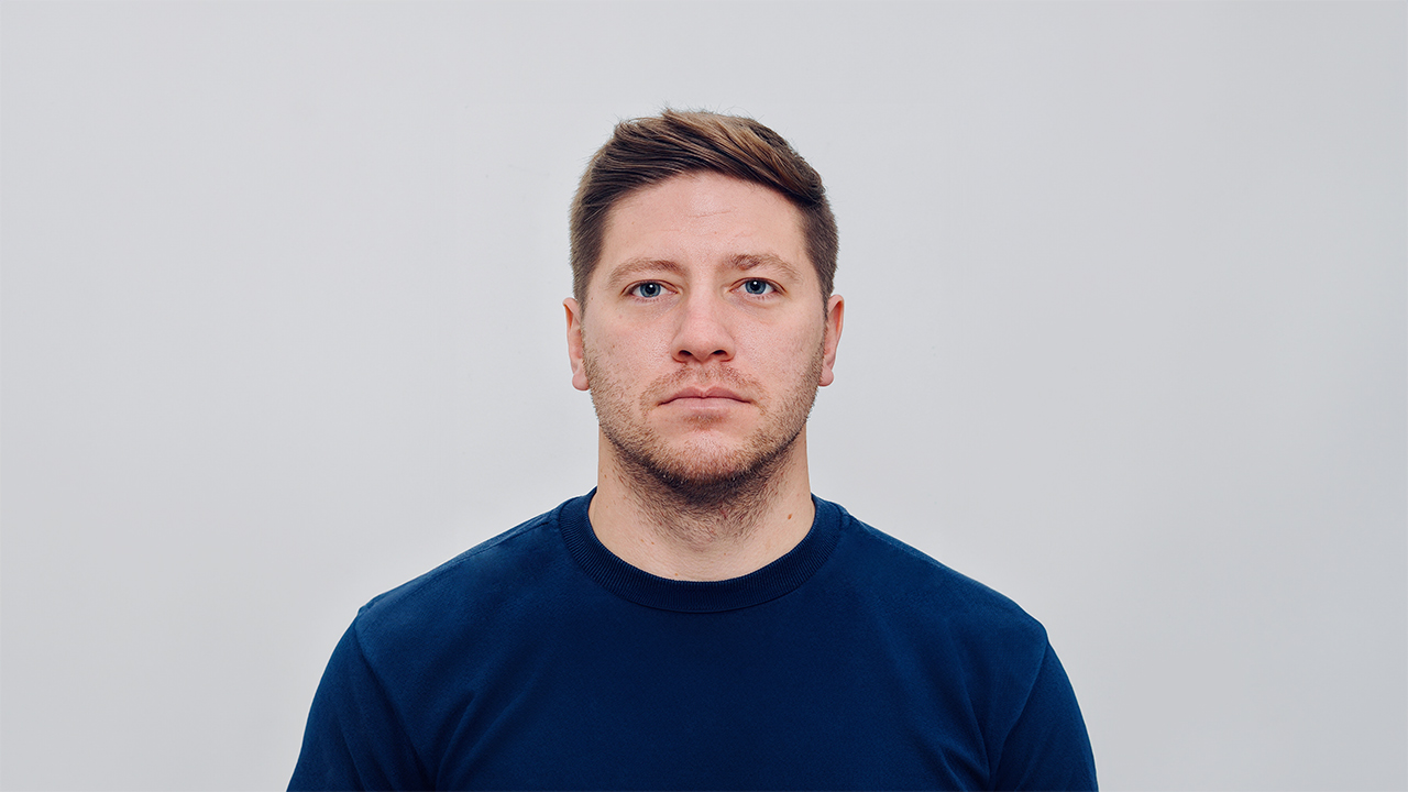 Headshot of George Firth - A man with short hair and a light beard is looking directly at the camera, wearing a navy blue t-shirt. The background is a plain light gray.