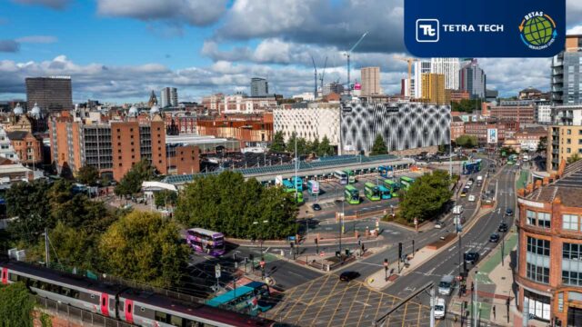 Aerial view of a busy urban area featuring a bus station with multiple green and blue buses, surrounding buildings, and a train passing through. The skyline includes a mix of modern and historical architecture under a partly cloudy sky.