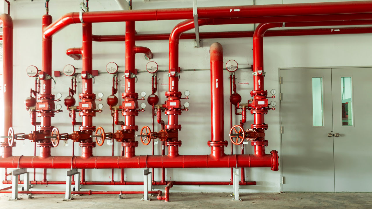 Red piping and valves and pressure gauges installed on a wall in an industrial setting.
