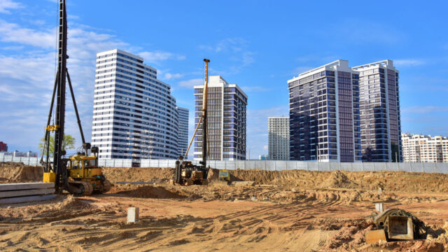 Construction site with drilling equipment in the foreground and modern residential buildings in the background.