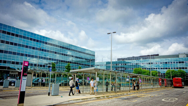 Covered bus stop with pedestrians in front of two blue buildings reflecting clouds