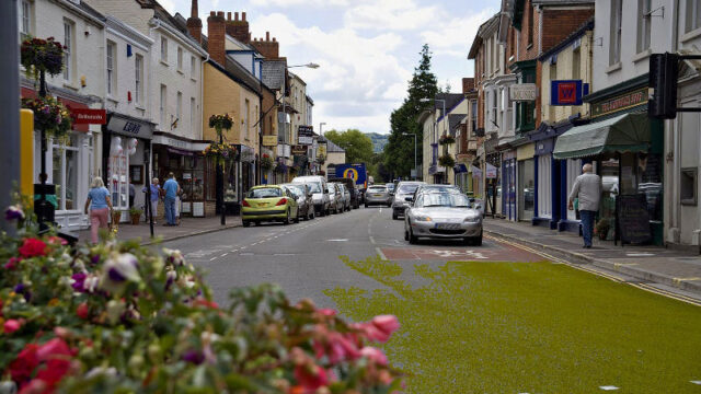 Busy street with parked cars, pedestrians, and green foliage