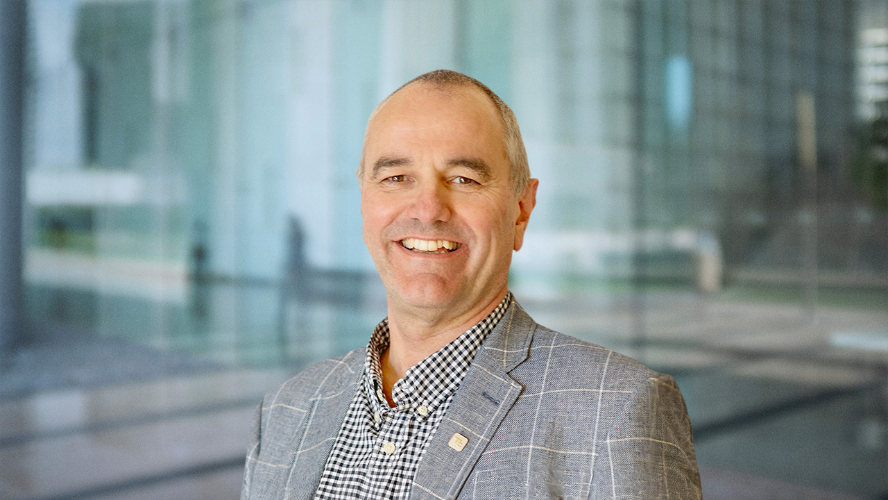 Headshot of Andrew Parkman - A man with short gray hair smiles at the camera, wearing a gray blazer over a checkered shirt. He stands in front of a glass wall, with a blurred background.