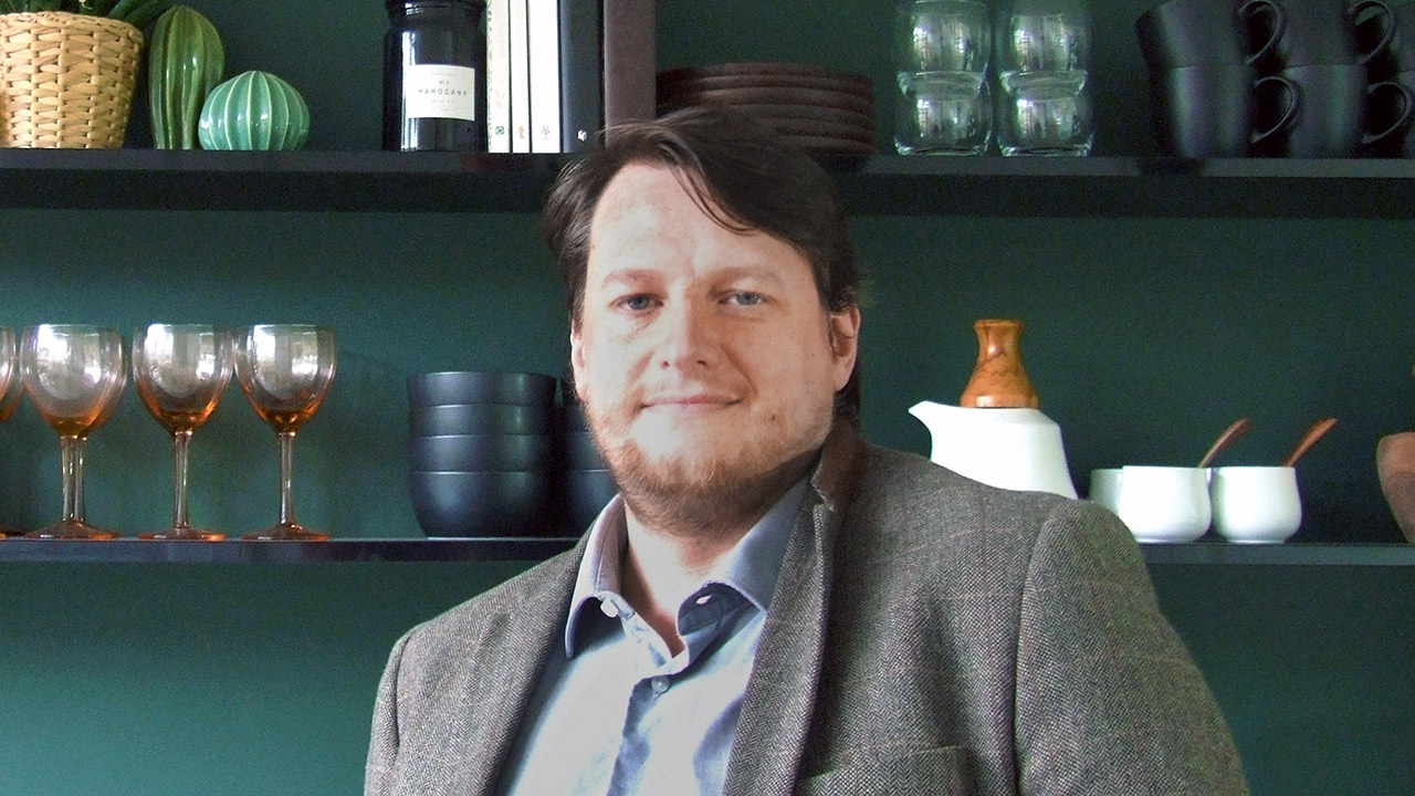 Headshot of Anthony Sherriff - A man with medium-length dark hair and a beard smiles at the camera. He is wearing a gray blazer over a light blue shirt, standing in front of a dark green wall with shelves displaying various kitchen items.