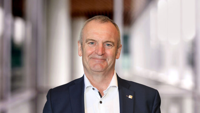 Headshot of Craig Hatch - A close-up portrait of a man with short hair and a slight smile, wearing a dark suit over a white shirt. The background is softly blurred, suggesting a modern indoor setting.