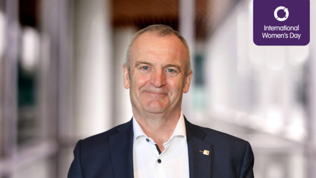 Headshot of Craig Hatch - A man in a suit is smiling at the camera, with a blurred background and a graphic indicating International Women’s Day in the corner.