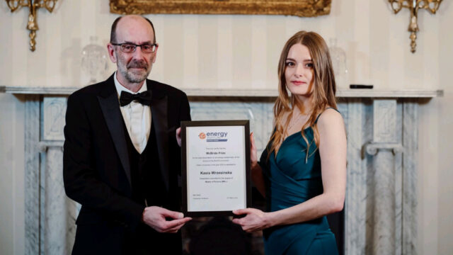 A man and a woman are holding a framed award certificate in a formal setting, with a decorative fireplace in the background.