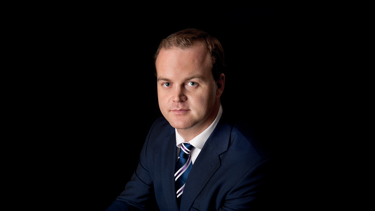 Headshot of Steven Thomas - A man with short hair is looking directly at the camera, wearing a dark suit and a striped tie. The background is black.