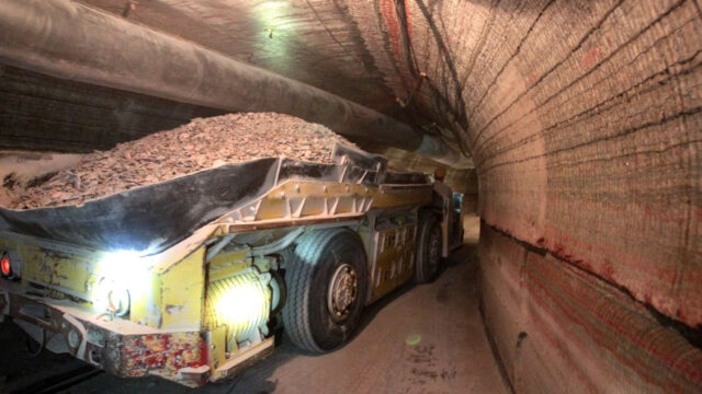 A mining truck loaded with material is navigating through a tunnel. The tunnel walls are lined with layers of rock and dust.