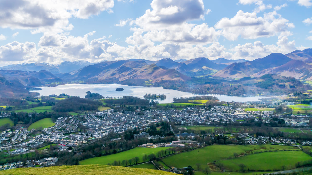 A panoramic view of a lake surrounded by mountains and greenery, with a small town visible in the foreground.