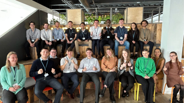 A group of young professionals is posing for a photo, smiling and giving thumbs up, in a modern office setting.