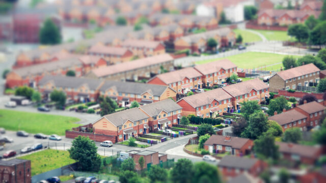 Aerial view of a housing development by Great Places Housing Group in north of England featuring rows of houses with red roofs and green lawns. The image has a tilt-shift effect, making it appear miniature.