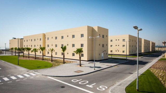 A view of a modern residential complex with beige buildings and palm trees, set against a clear blue sky. The street features a "STOP" sign.