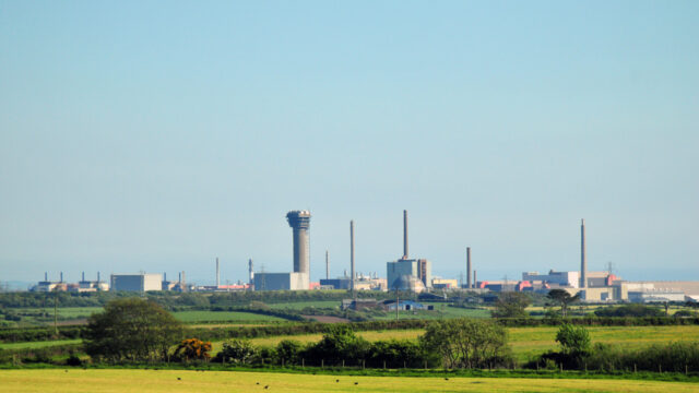A distant view of an industrial complex with several smokestacks and structures, set against a clear blue sky and green fields.