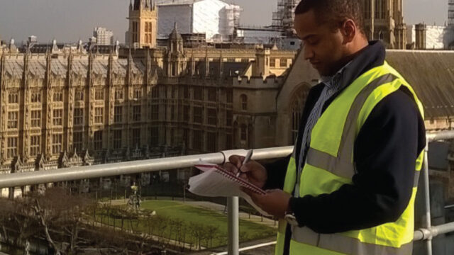 A man wearing a high-visibility vest is standing on a balcony, taking notes in a notebook. The background features the intricate architecture of a historic building, likely the Houses of Parliament, with scaffolding visible.