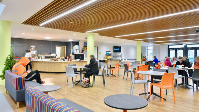 Interior view of the sports hall lobby with a cafe bar and several tables with people lounging or enjoying their food