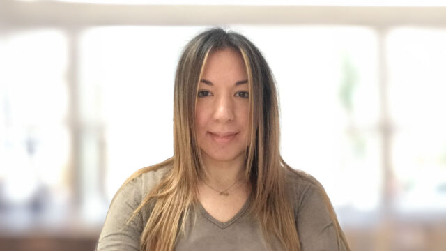 A woman, Eleftheria Mazioti, with long, straight hair and a neutral expression is facing the camera. She is wearing a gray top, and the background is softly blurred with bright light.