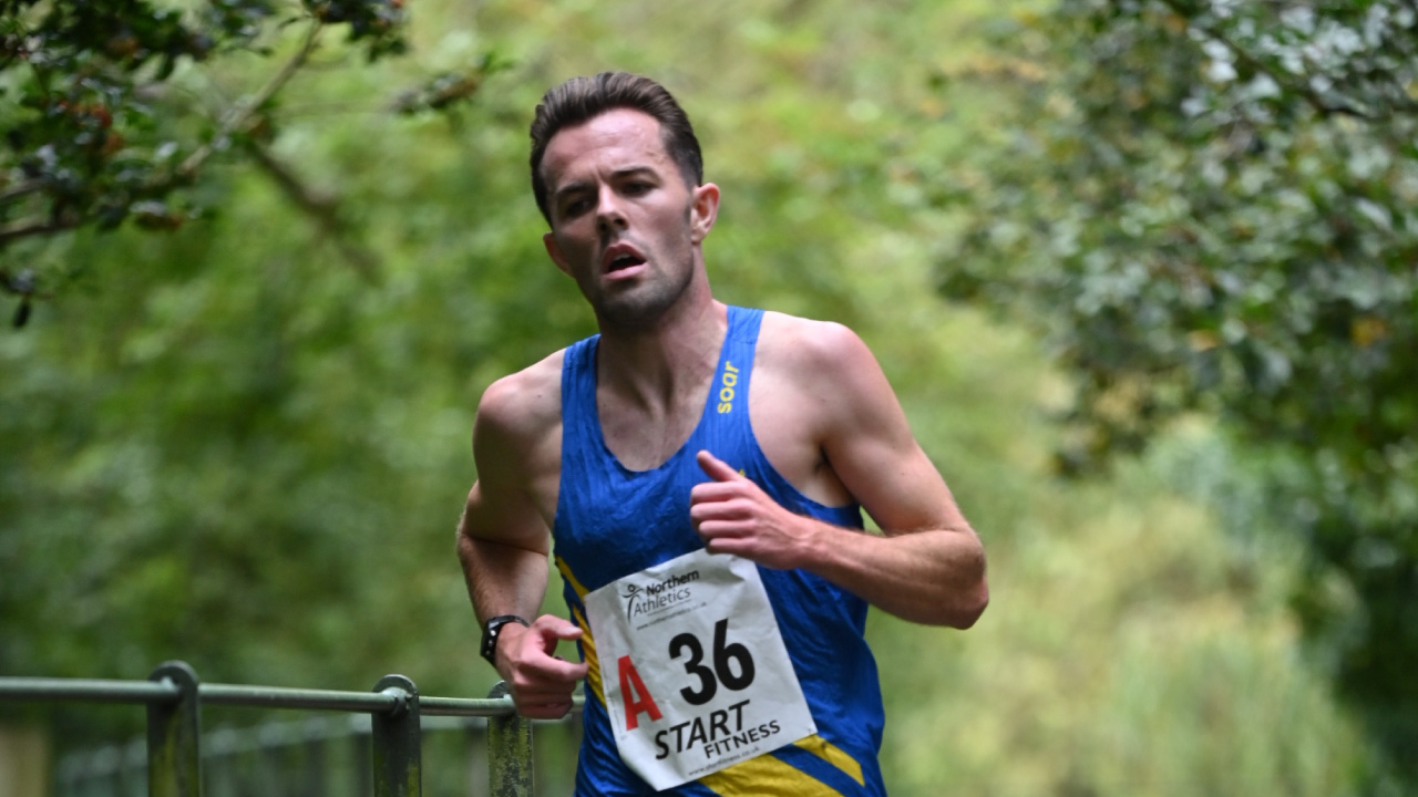 A male runner is competing in a race, wearing a blue tank top with yellow accents and a race number pinned to his chest. He appears focused and is running along a tree-lined path.