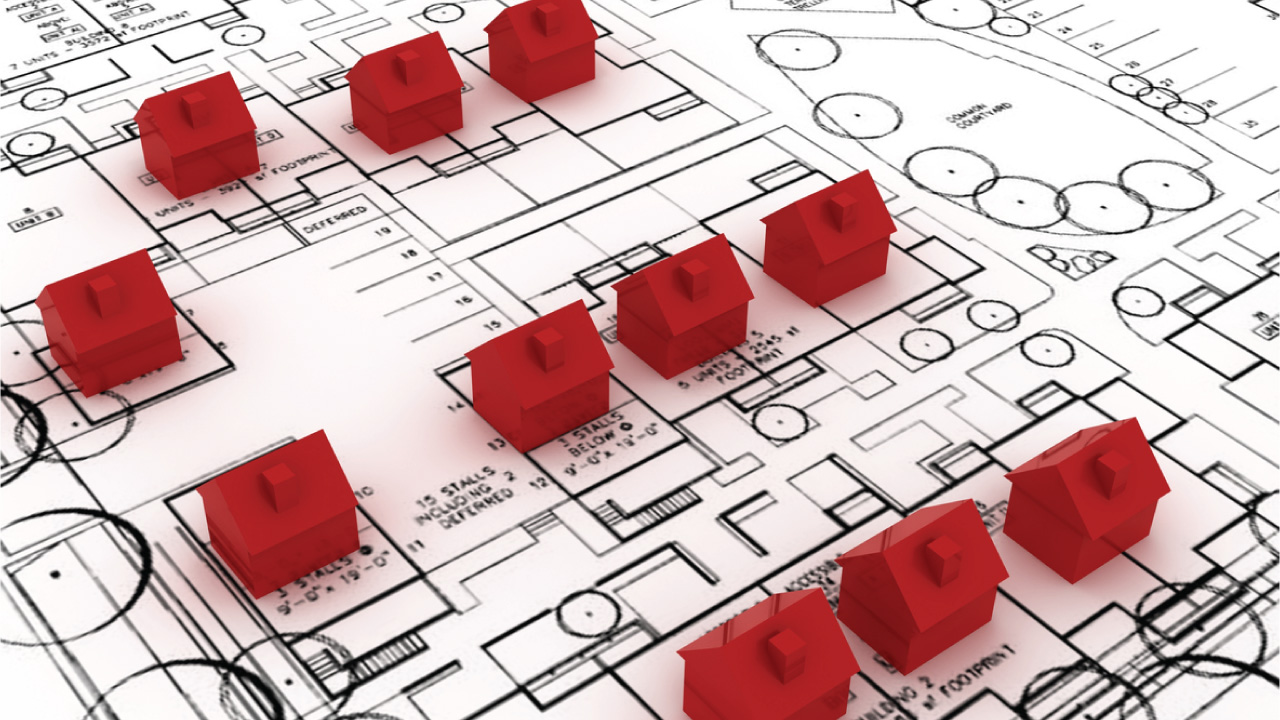 A top-down view of a building blueprint with several red house-shaped models placed on it. The blueprint features detailed architectural plans, including rooms and layout designs.