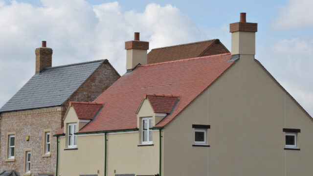 A close-up view of two houses with different roof styles. The house on the left has a slate roof, while the house on the right features a red tiled roof. Both houses have brick chimneys.