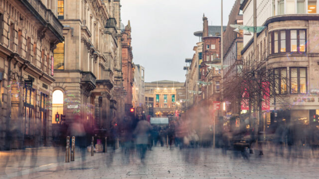 A busy urban street scene with blurred figures of pedestrians walking along the pavement. The background features historic buildings and a large structure, likely a shopping center, illuminated in the distance.