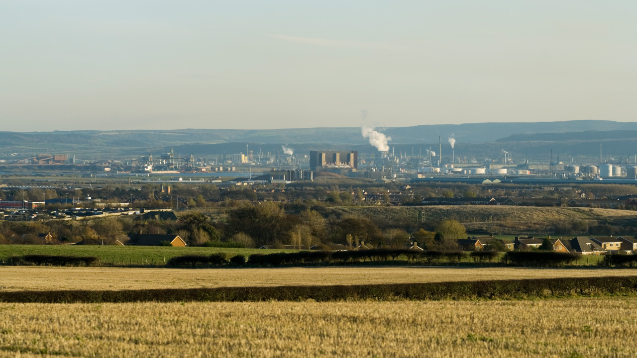 A panoramic view of an industrial landscape with factories and smokestacks in the distance, surrounded by fields and residential areas.