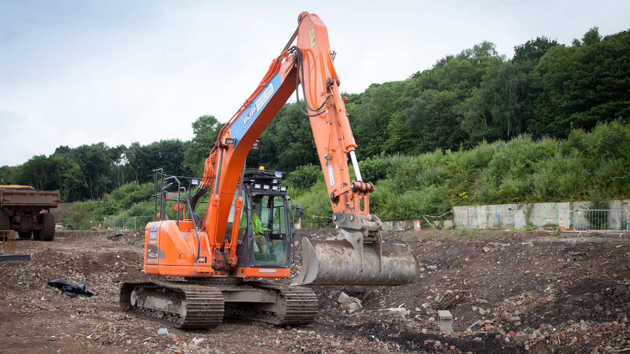 An orange excavator on a construction site, with a green operator visible inside the cabin, working on a rocky terrain.