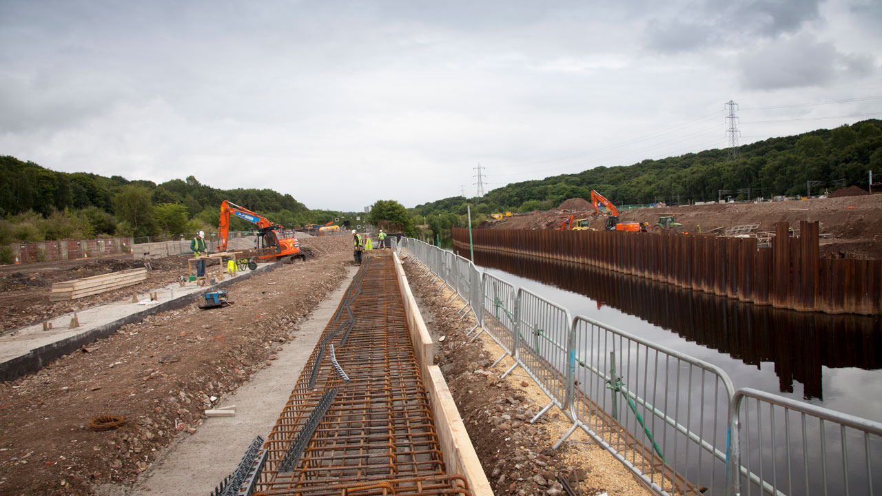 A construction site along a river, featuring excavators, workers, and rebar structures, with a wooded area in the background.