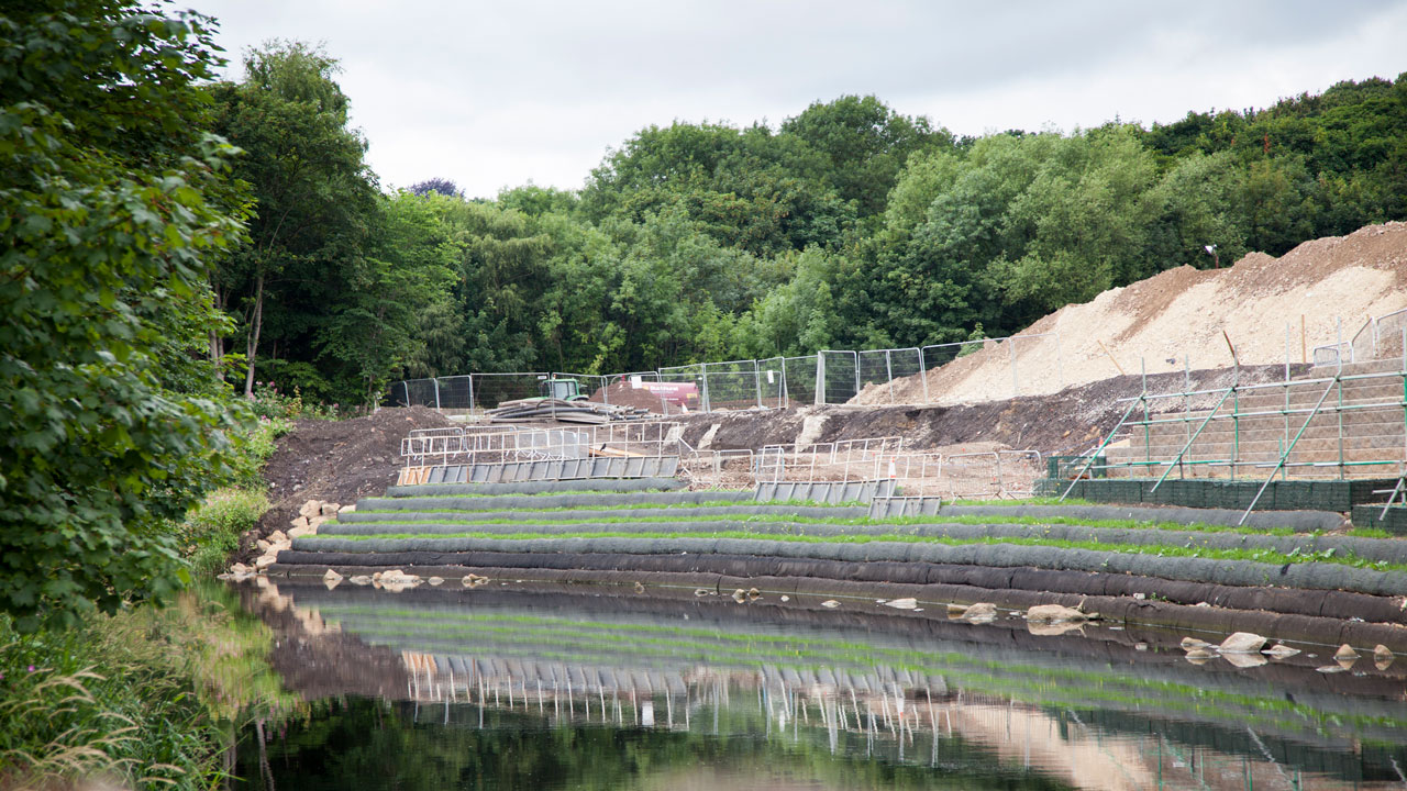 A construction site along a riverbank, featuring terraced earthworks, fencing, and greenery in the background.