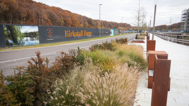 Work area advertisement banner for Kirkstall Forge along the side of road running by orange-leafed forest edge near a city walkway