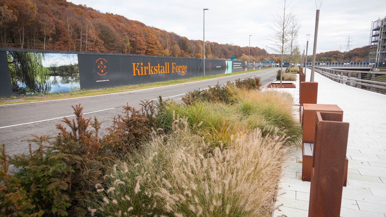 A landscaped area along a road featuring decorative grasses, a sign for "Kirkstall Forge," and a backdrop of autumn trees.