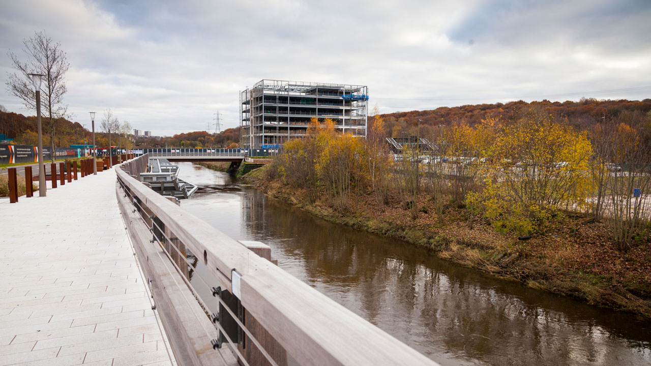 A riverside walkway with a wooden railing, overlooking a construction site and autumn foliage along the water.
