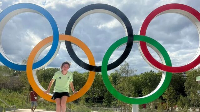 Clara Evans Stood in front of an Olympics 2024 Paris statue - A young woman poses playfully between the Olympic rings, smiling at the camera.