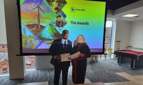 Two individuals stand together holding a certificate in front of a presentation screen displaying the Tetra Tech logo and the title "The Awards."