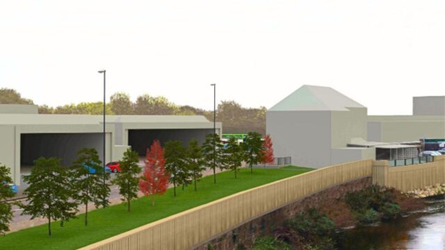 An architectural rendering of a development area near a river. The image features a row of trees, some with red foliage, along a grassy area adjacent to a building with large openings. A wooden fence runs along the riverbank, and several vehicles are parked nearby. The background includes trees and a clear sky.