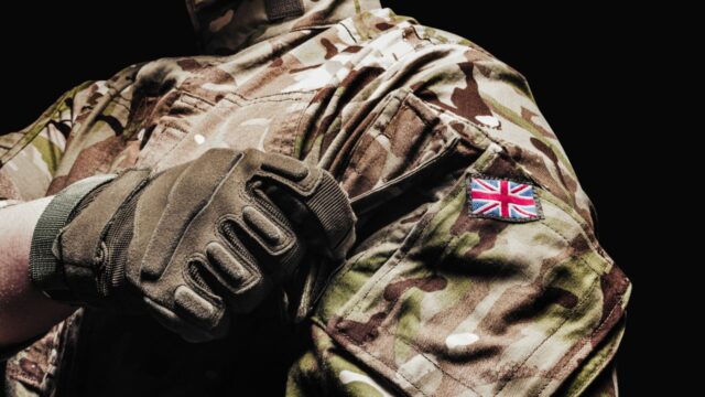 Close-up of a soldier's arm in camouflage uniform with a British flag patch, wearing a tactical glove, against a black background.