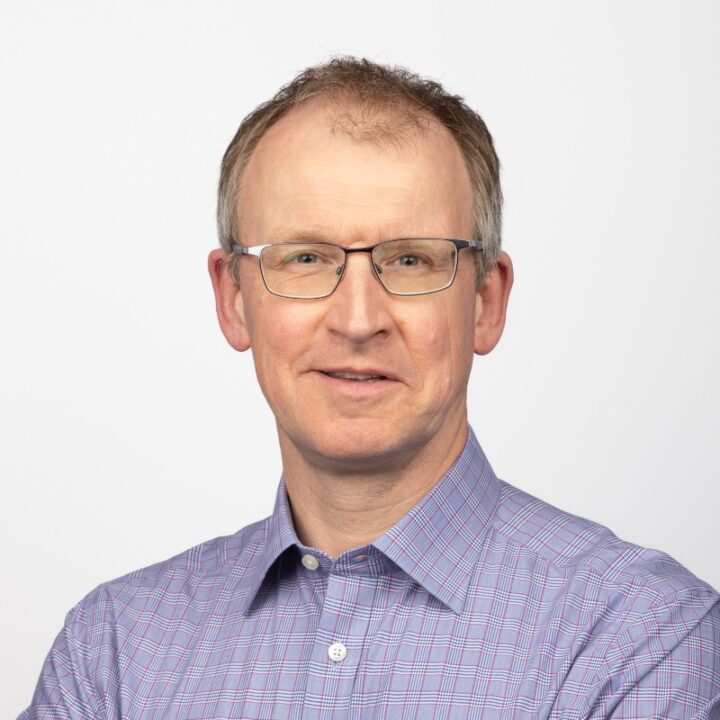 Middle-aged man with short light brown hair and glasses, wearing a purple checkered shirt, smiling slightly against a plain white background.