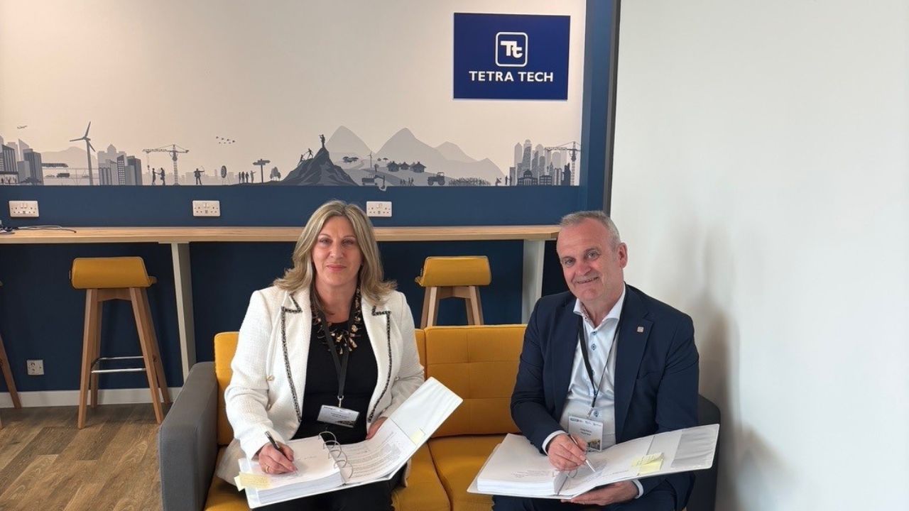 Two professionals, a woman in a white blazer and a man in a dark suit, sitting on a yellow and gray sofa signing large documents, with a blue wall behind them featuring a cityscape mural and the Tetra Tech logo.
