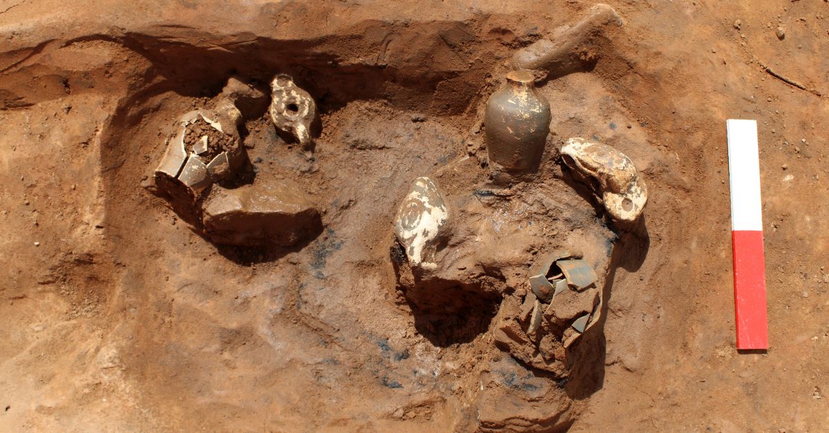 An archaeological excavation pit containing several ancient pottery fragments and a small intact vessel, with a red and white measuring scale placed on the right side.