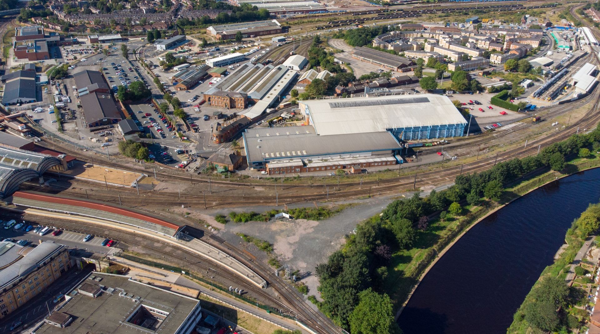 Aerial view of a large railway yard with multiple tracks, industrial buildings, parked cars, and a river curving along one side of the scene.