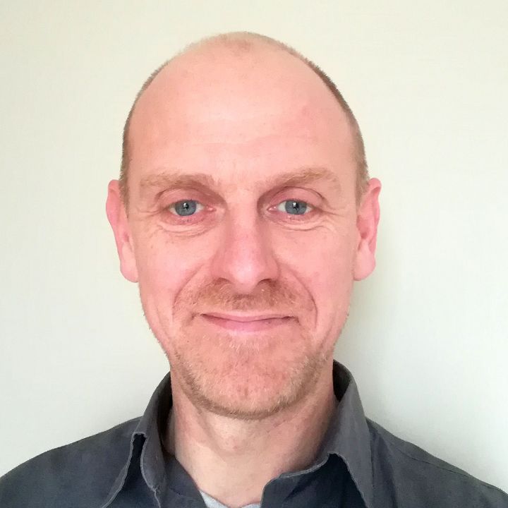 A close-up portrait of a middle-aged man with blue eyes, a light beard, and a receding hairline, smiling gently while wearing a dark gray collared shirt.
