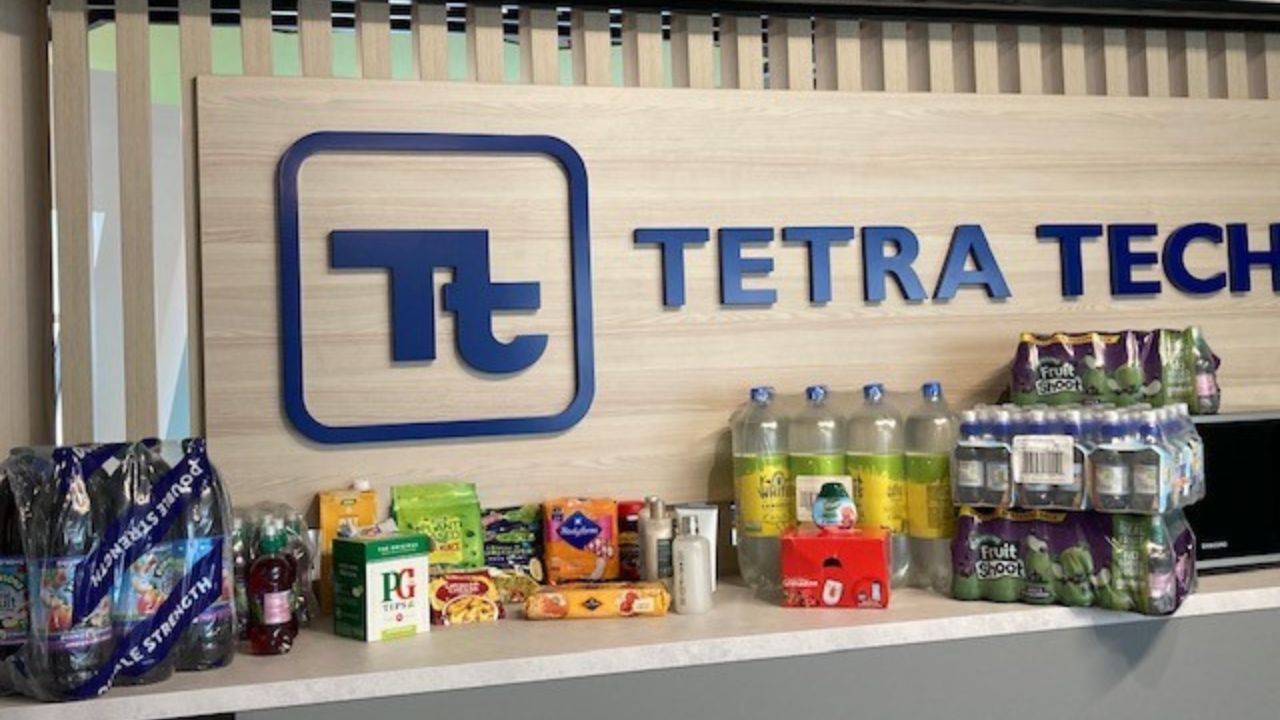 A counter in front of a wooden wall with a blue "Tetra Tech" logo, displaying various packaged food and drink items including bottled water, fruit drinks, tea, biscuits, and snacks