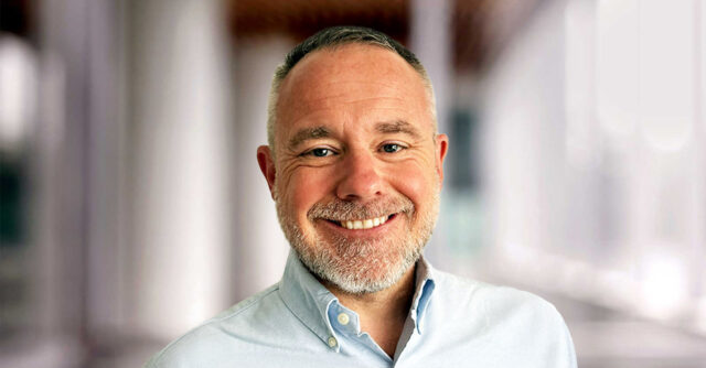 Portrait of Kev Thomas smiling he is a man with short gray hair and a beard, wearing a light blue button-up shirt, standing in a softly blurred indoor setting.