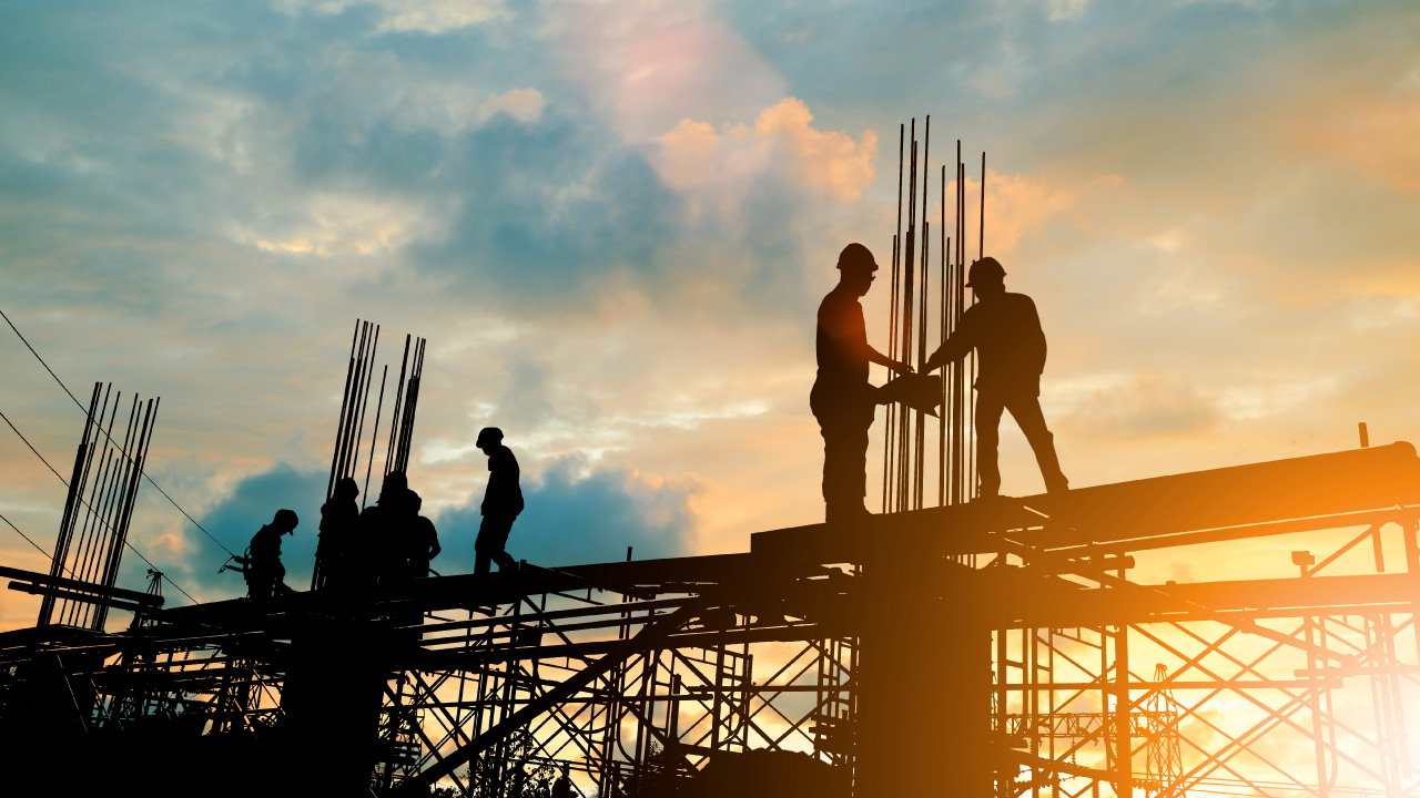 Silhouetted construction workers on a steel framework installing vertical rebar against a golden sunset sky.
