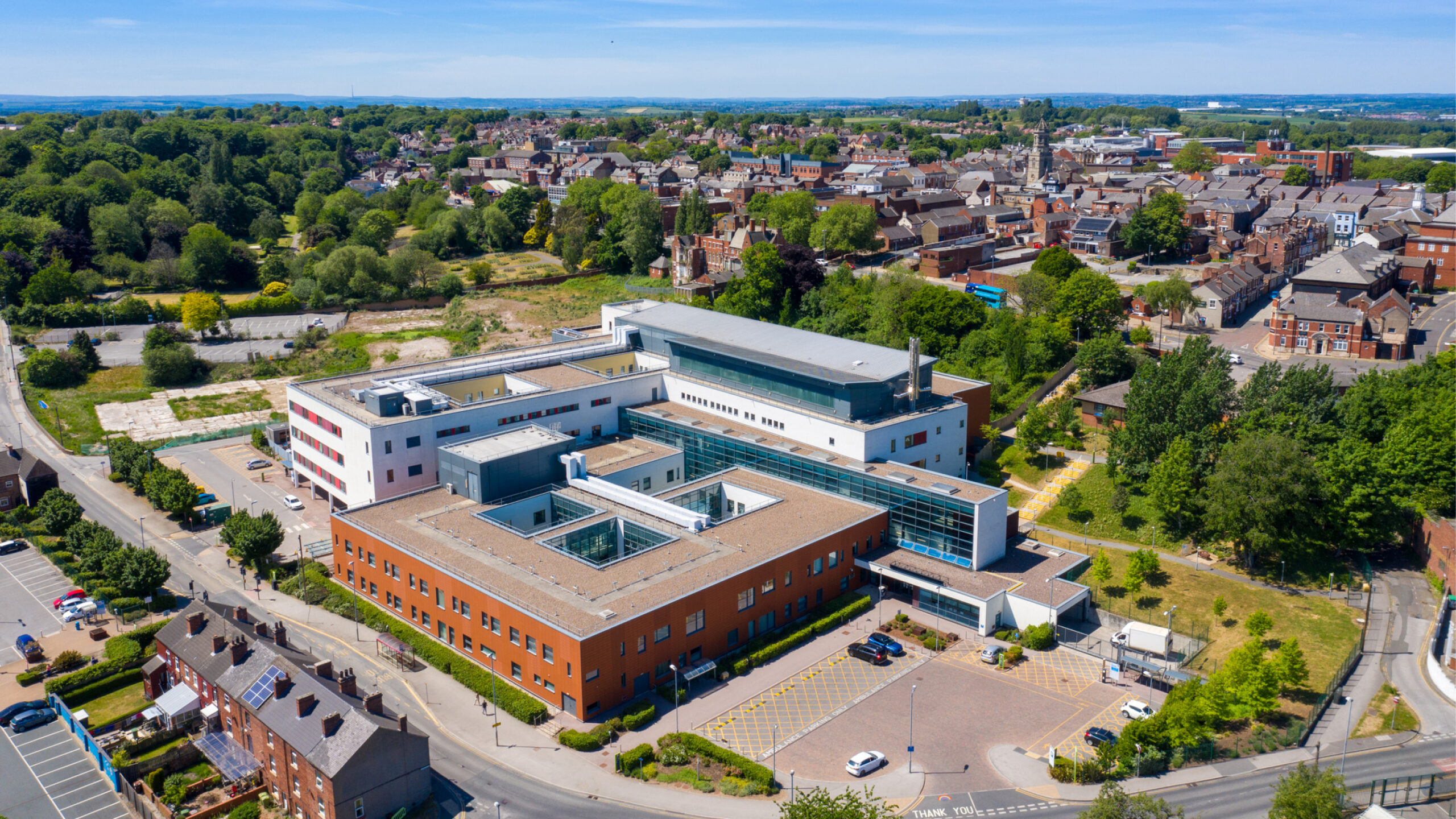 Aerial view of a modern multi-wing civic building and surrounding parking, with residential streets and a tree-filled park in the background.
