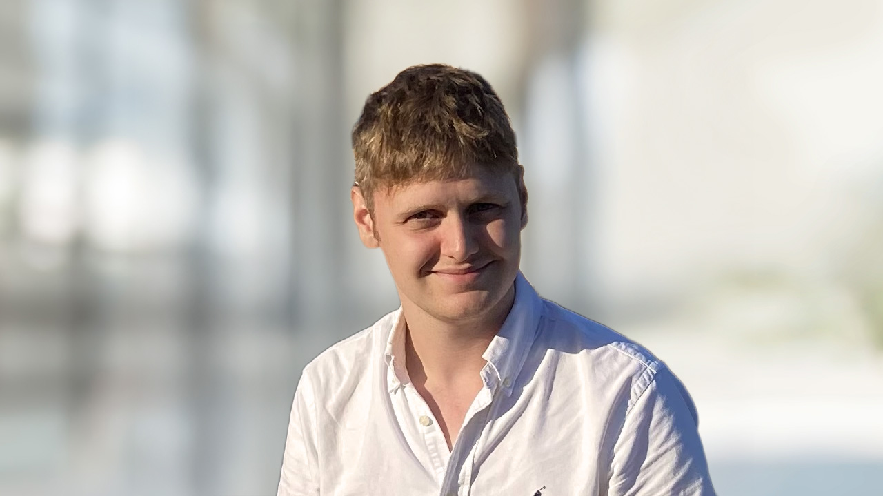 Young man with short light-brown hair wearing a white button-up shirt, smiling against a softly blurred indoor background.