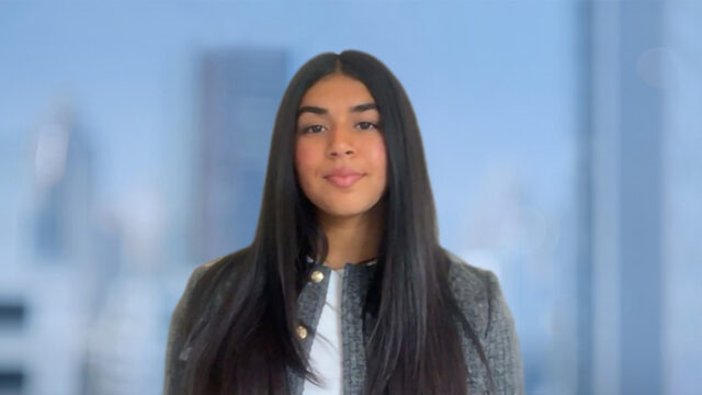 Young woman with long dark hair wearing a gray jacket, facing camera against a softly blurred blue cityscape background.